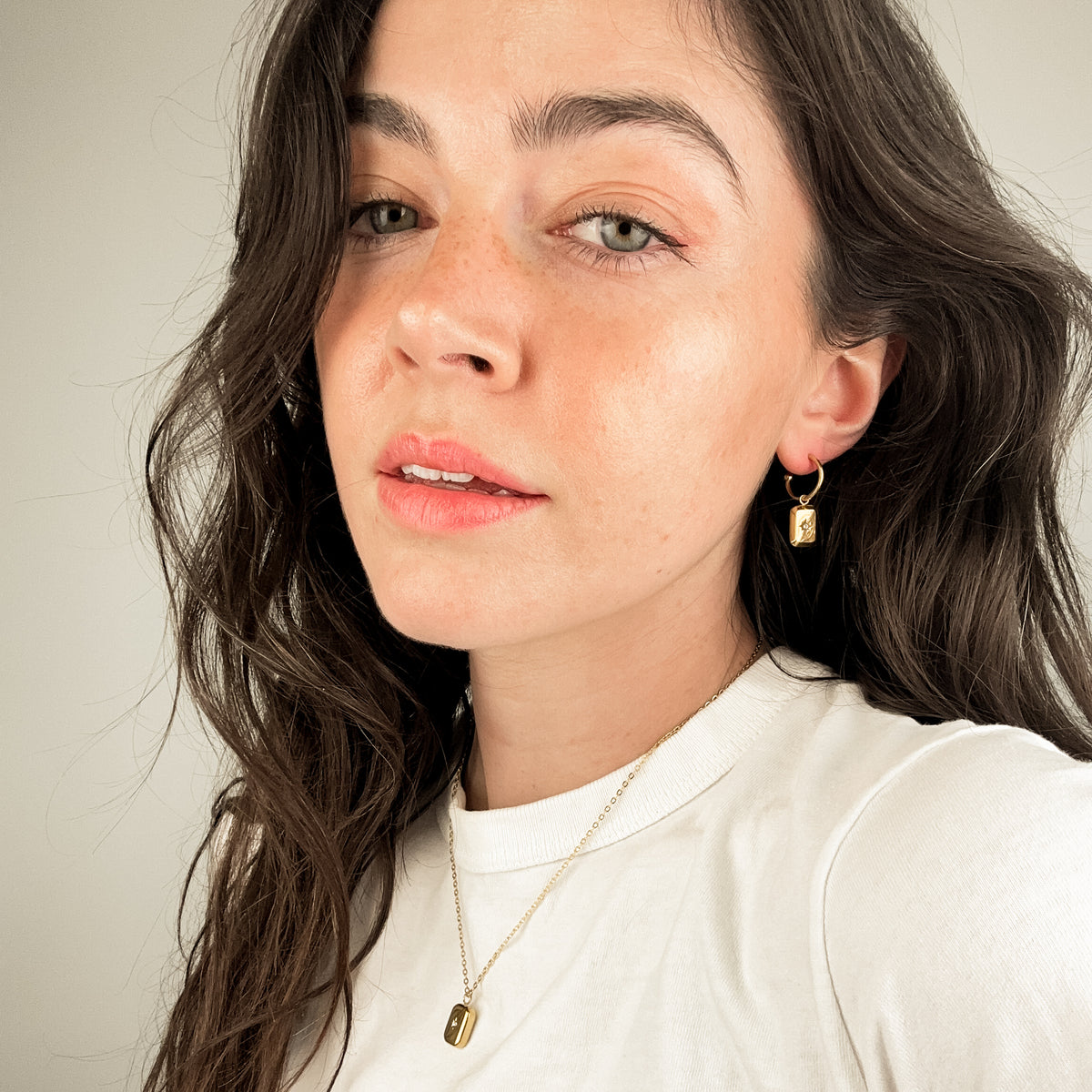 model is wearing gold earrings with a delicate dangling charm. the charm can be removed also and the earrings can be worn as simple hoops. The charm that hangs on the hoop is embellished with a star. the model is also wearing the matching poise necklace. 