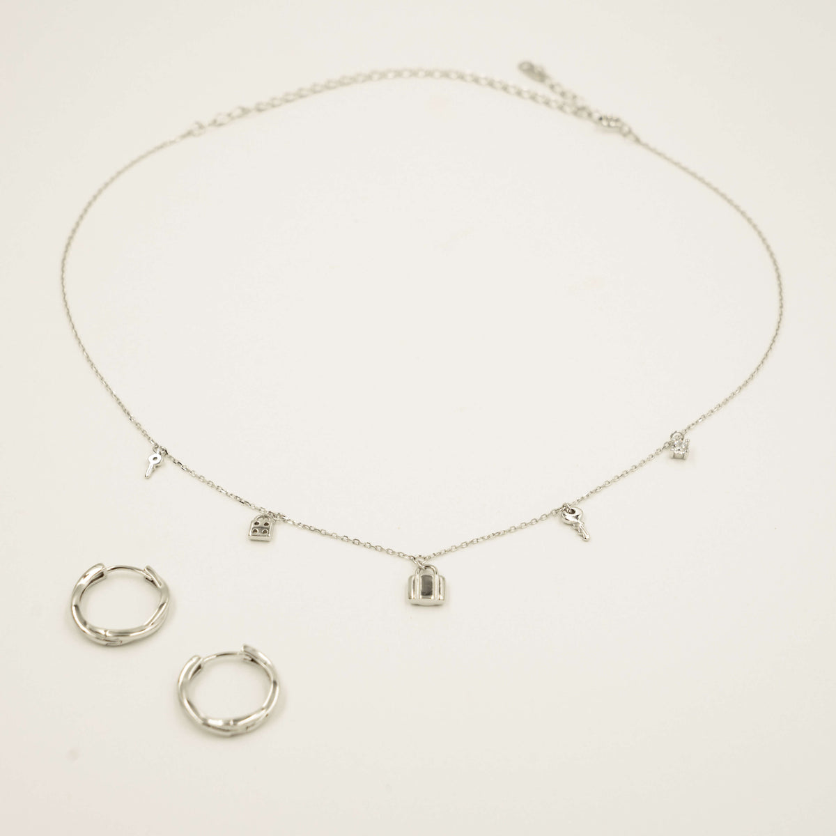 Earrings and a necklace together. The earrings are hoop earrings. The charm necklace has five silver charms on a silver chain. The charms include two a diamonds, two keys and a suitcase.