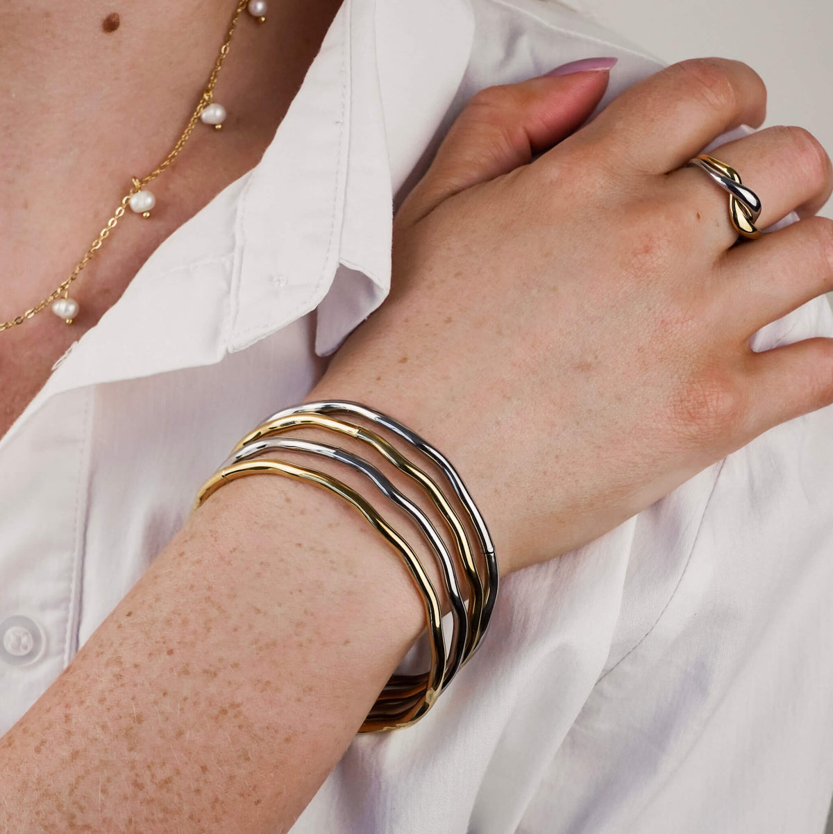 waterproof stacking bangles in silver and gold. The model is wearing four lumina bangles stacked together and she is also wearing the matching lumina two tone ring which is also from Mettle & Bloom. She is holding her hand to her shoulder, against her white shirt. 