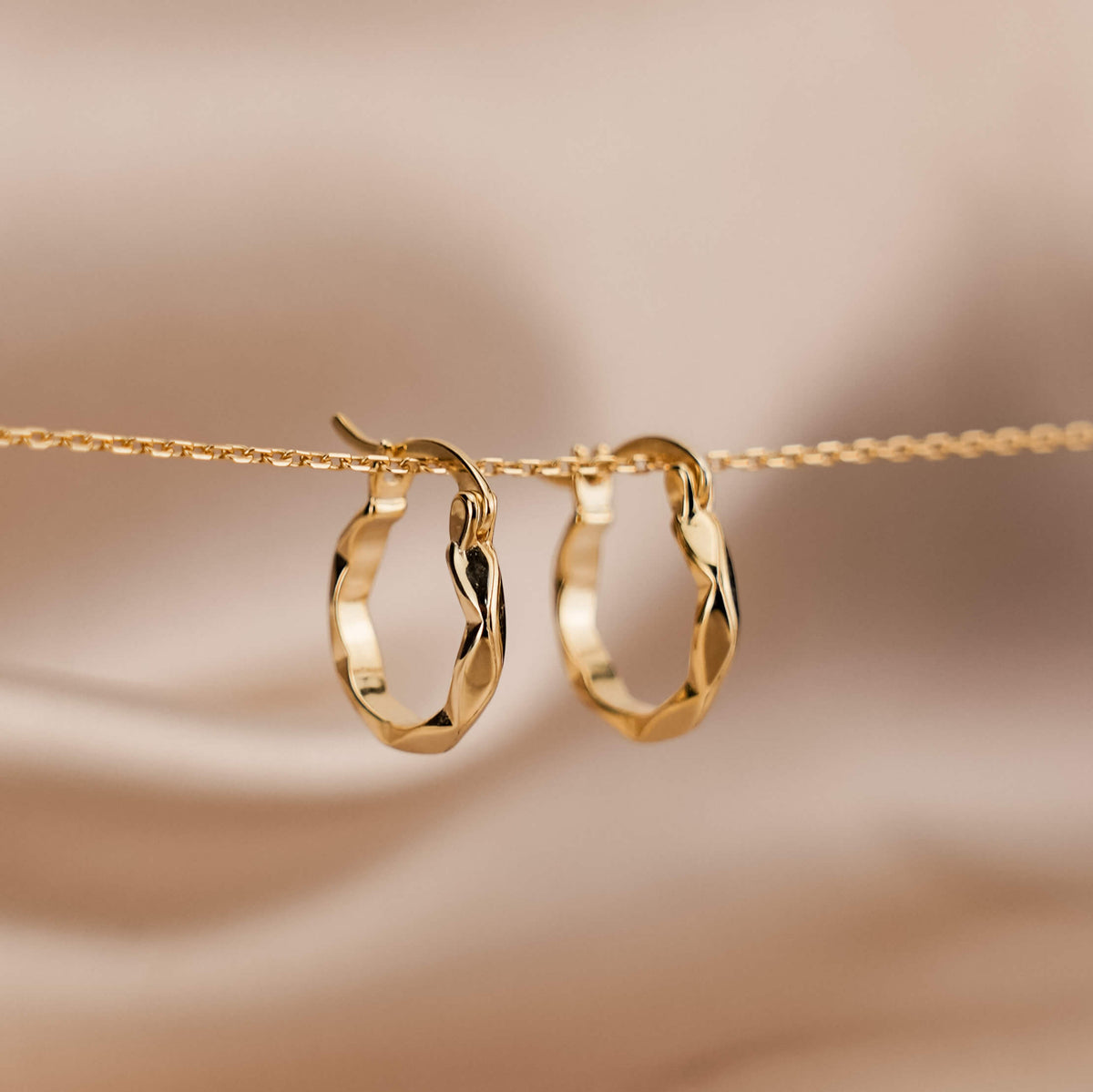 Gold hoop earrings made from high quality sterling silver plated in 18 karat gold. They have a subtle twist design. The earrings are hanging from a fine, dainty gold chain in front of pink silk.