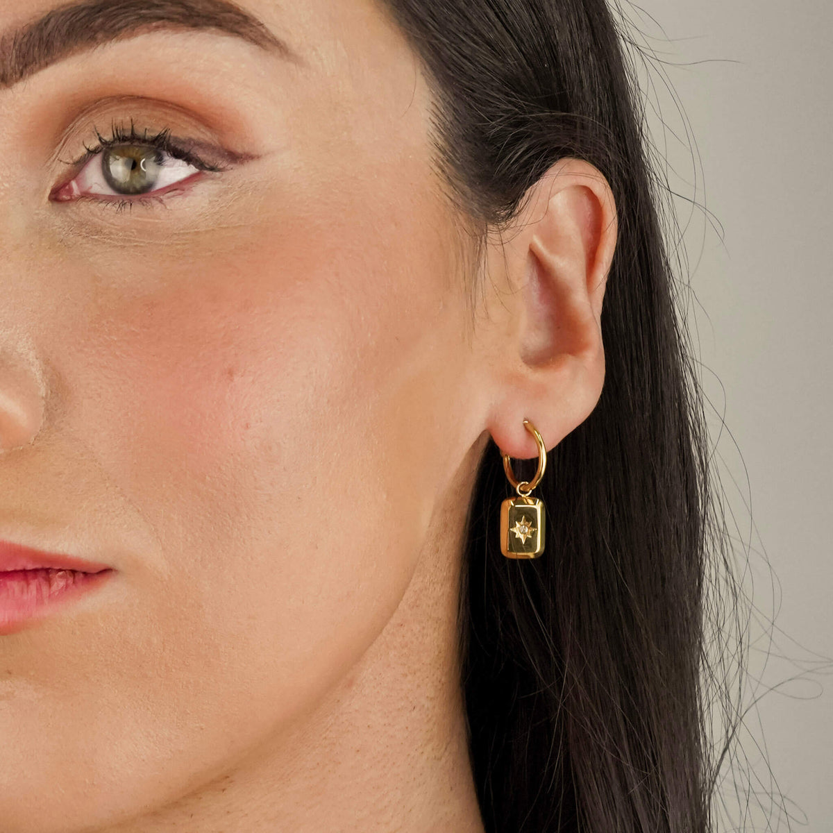 model is wearing gold earrings with a delicate dangling charm. the charm can be removed also and the earrings can be worn as simple hoops. The charm that hangs on the hoop is embellished with a star. 