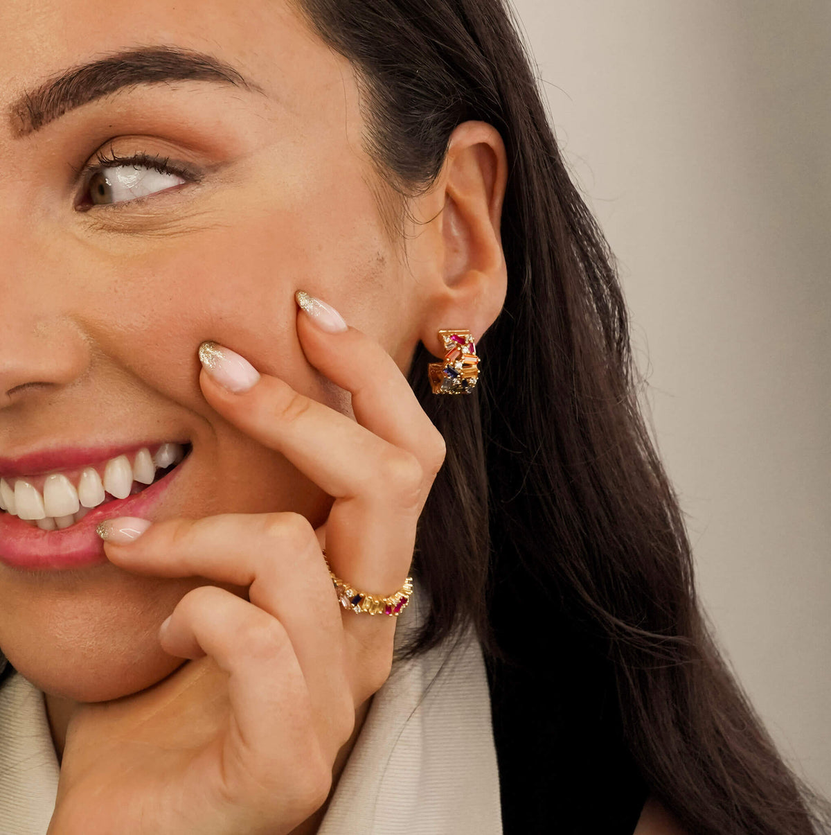 a model is wearing  the Multicoloured adorn ring from an Irish jewellery brand. The adorn ring is a colourful piece of jewellery. The sterling silver band is surrounded and covered in multicoloured zirconia stones which are set in unique and random ways.  The model is also wearing the matching Regalia hoop earrings. 