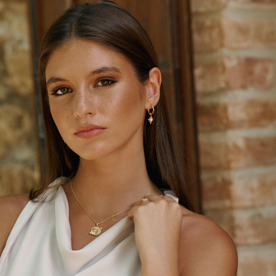 Woman wearing gold jewelry against a brick wall. they are the star drop earrings from mettle and bloom. they were designed in house by mettle and bloom. they are a hoop earring with a star suspended from it. She styled them with another custom design from Mettle and Bloom which is the undiscovered treasures necklace. 