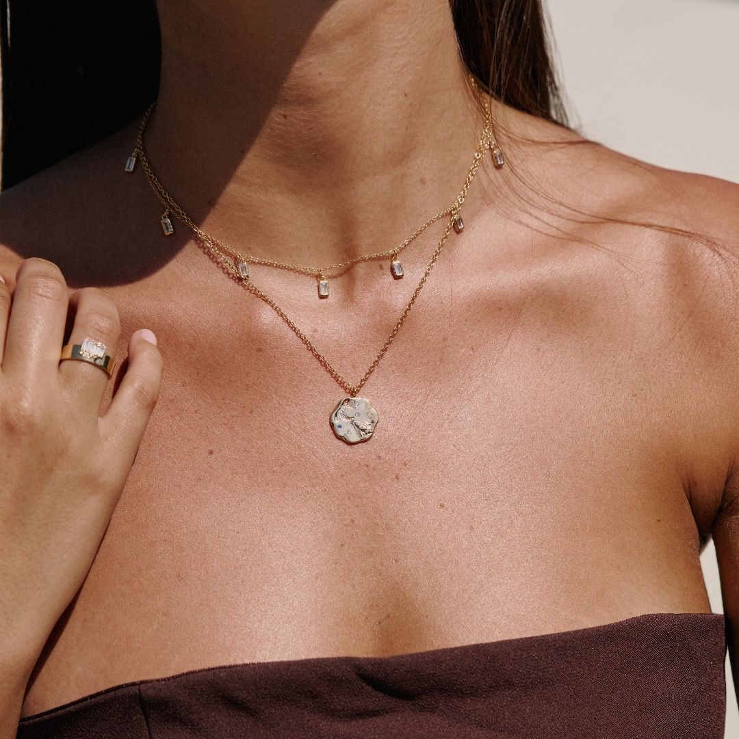 Close-up of a person wearing a gold necklace with a To the moon and back pendant and molten chakra necklace, with a blurred background. She is also wearing a mettle and bloom must have, the vega ring