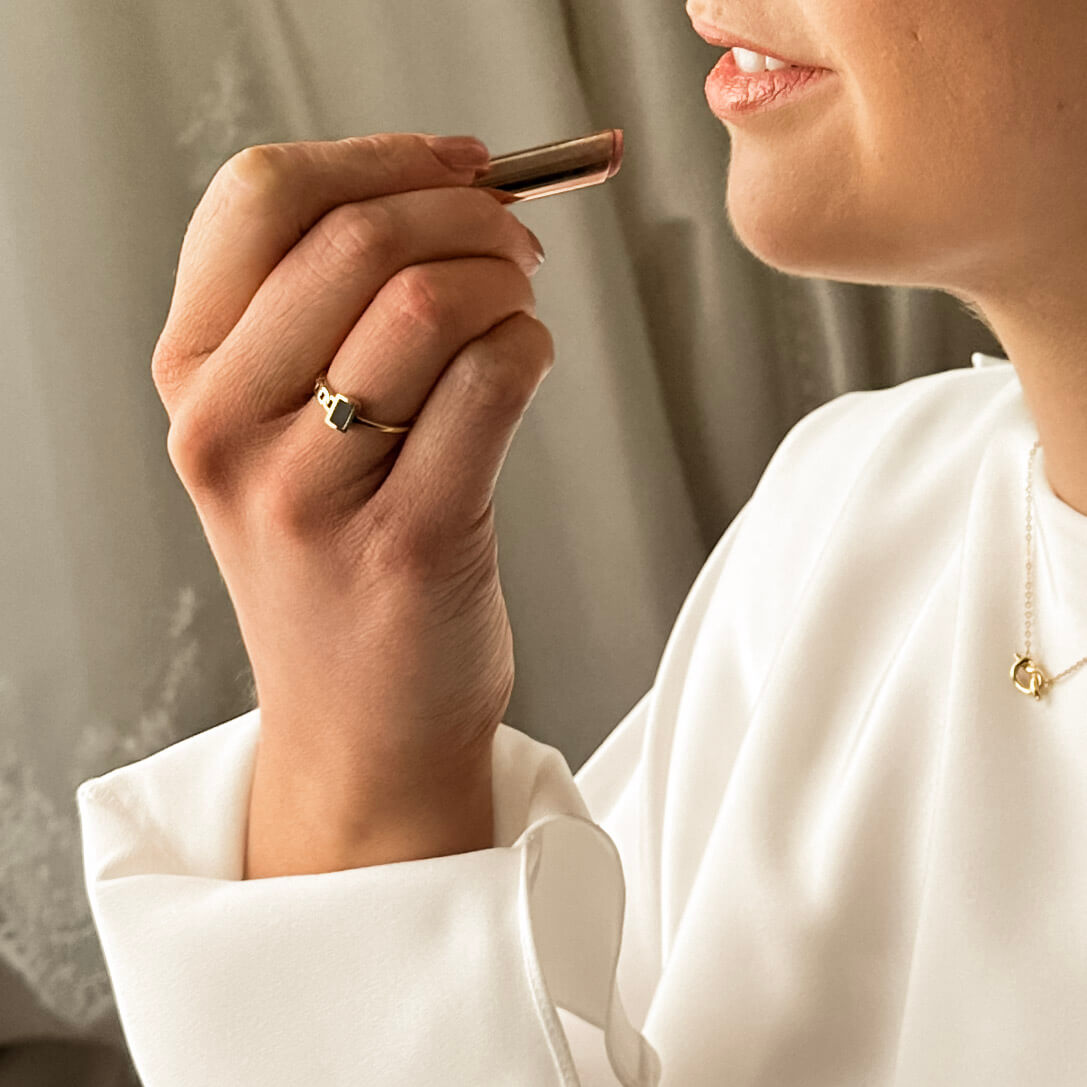 a bride is wearing a unique gold ring with a black stud on it's face while she applied her lipstick for her wedding day. 