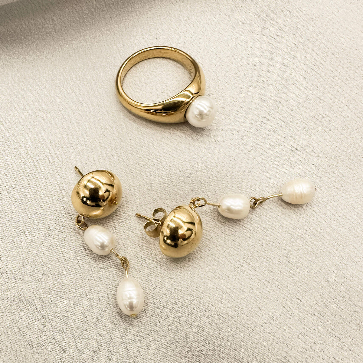 gold band ring with a pearl embedded in it is laid on it's side on a silk surface. It is beside the Adeline pearl earrings. 