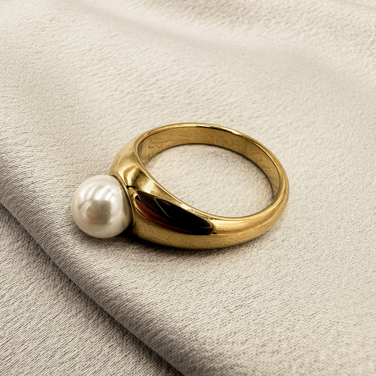 the amara ring is laid flat on a silk surface. the ring has a gold band that is chunky and holds a pearl. 
