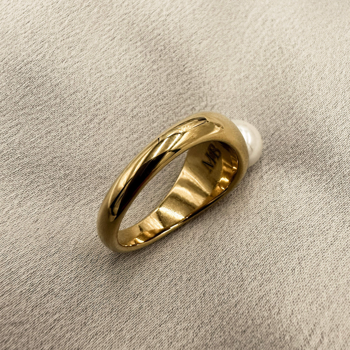 gold band ring with a pearl embedded in it is laid on it's side on a silk surface. You can see the M&B logo. 